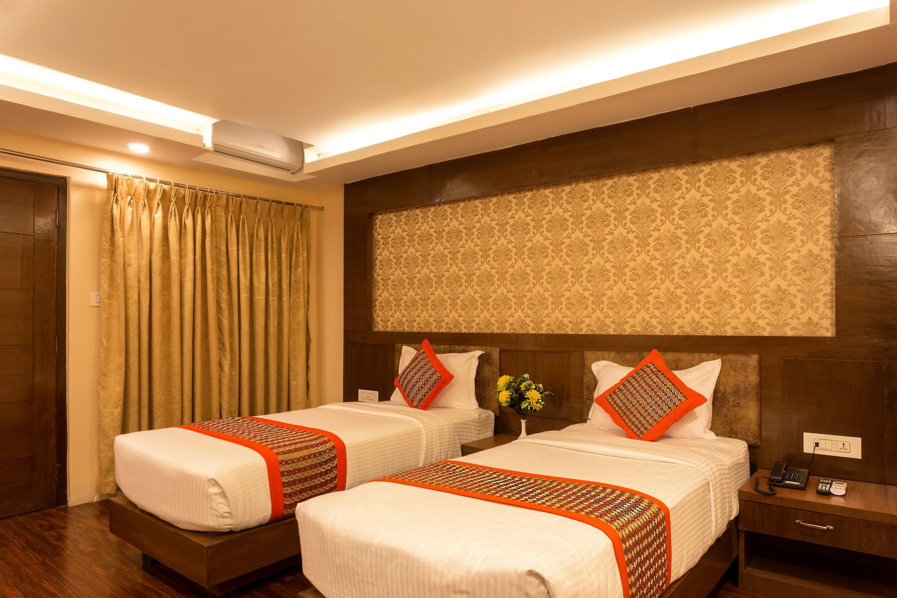 A hotel room with two beds, a wooden headboard, a nightstand with a phone, and a wall-mounted air conditioner.