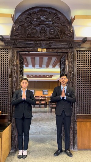 Two individuals in formal attire standing at a wooden entrance with intricate carvings, a chandelier, and a seating area visible in the background. - Hotel Yellow Pagoda Pokhara (Pokhara)