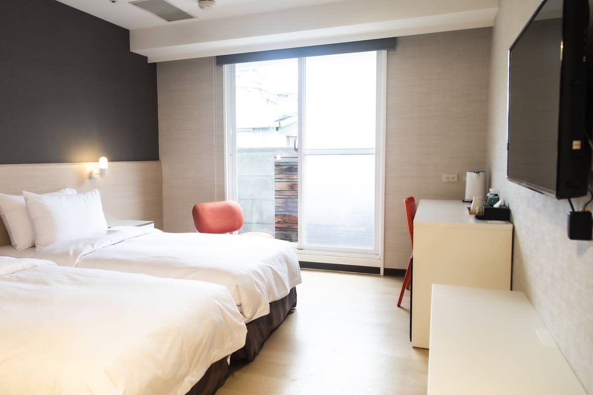 Une chambre d’hôtel moderne avec deux lits, un fauteuil rouge, un bureau avec une machine à café et une grande fenêtre.