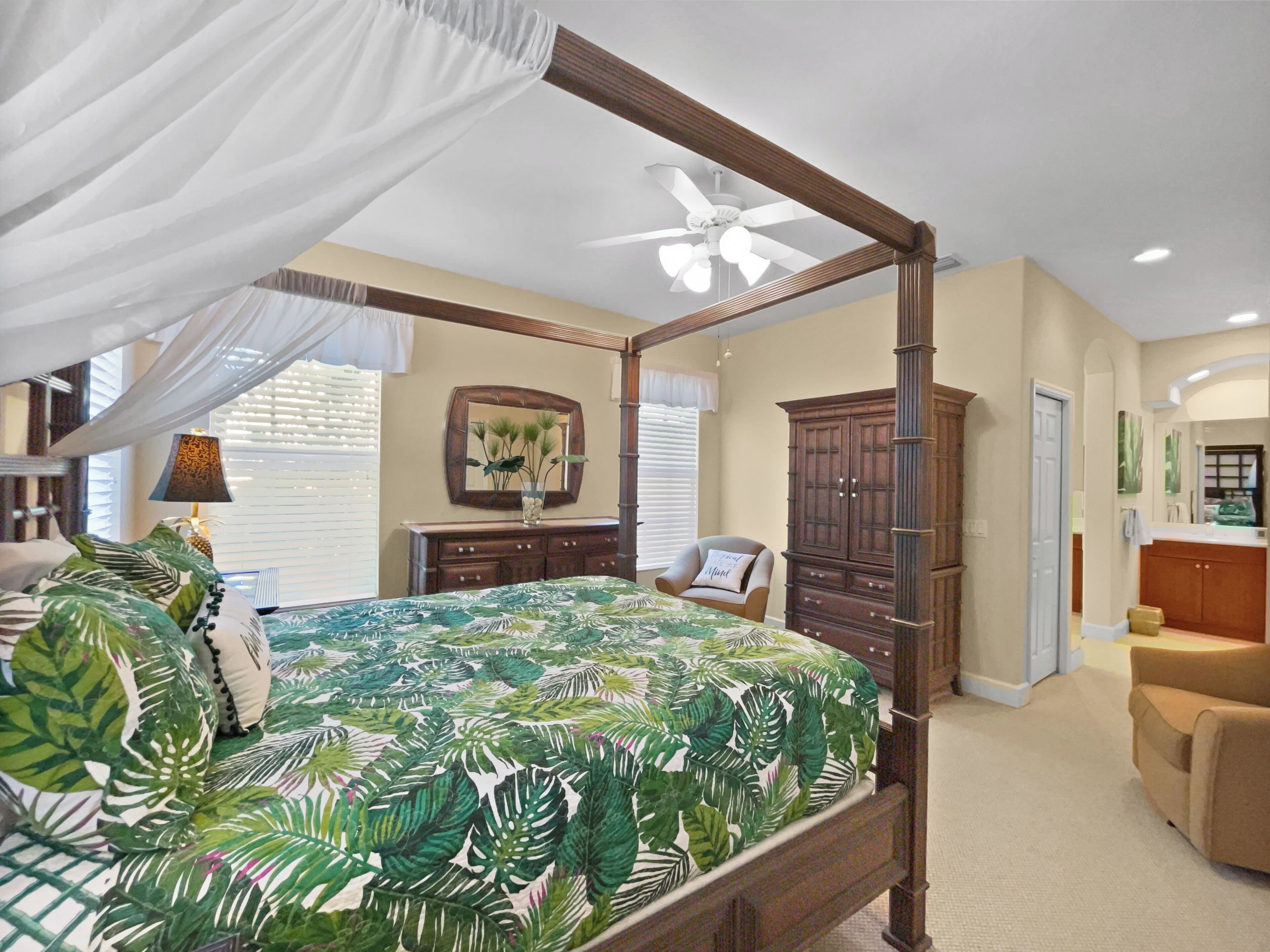 A four-poster bed with a tropical leaf bedspread, a wooden nightstand, a dresser, a chair, and a ceiling fan.