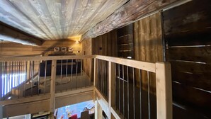 A rustic cabin interior with a wooden ceiling, a balcony with a railing, and a table with various items on it.