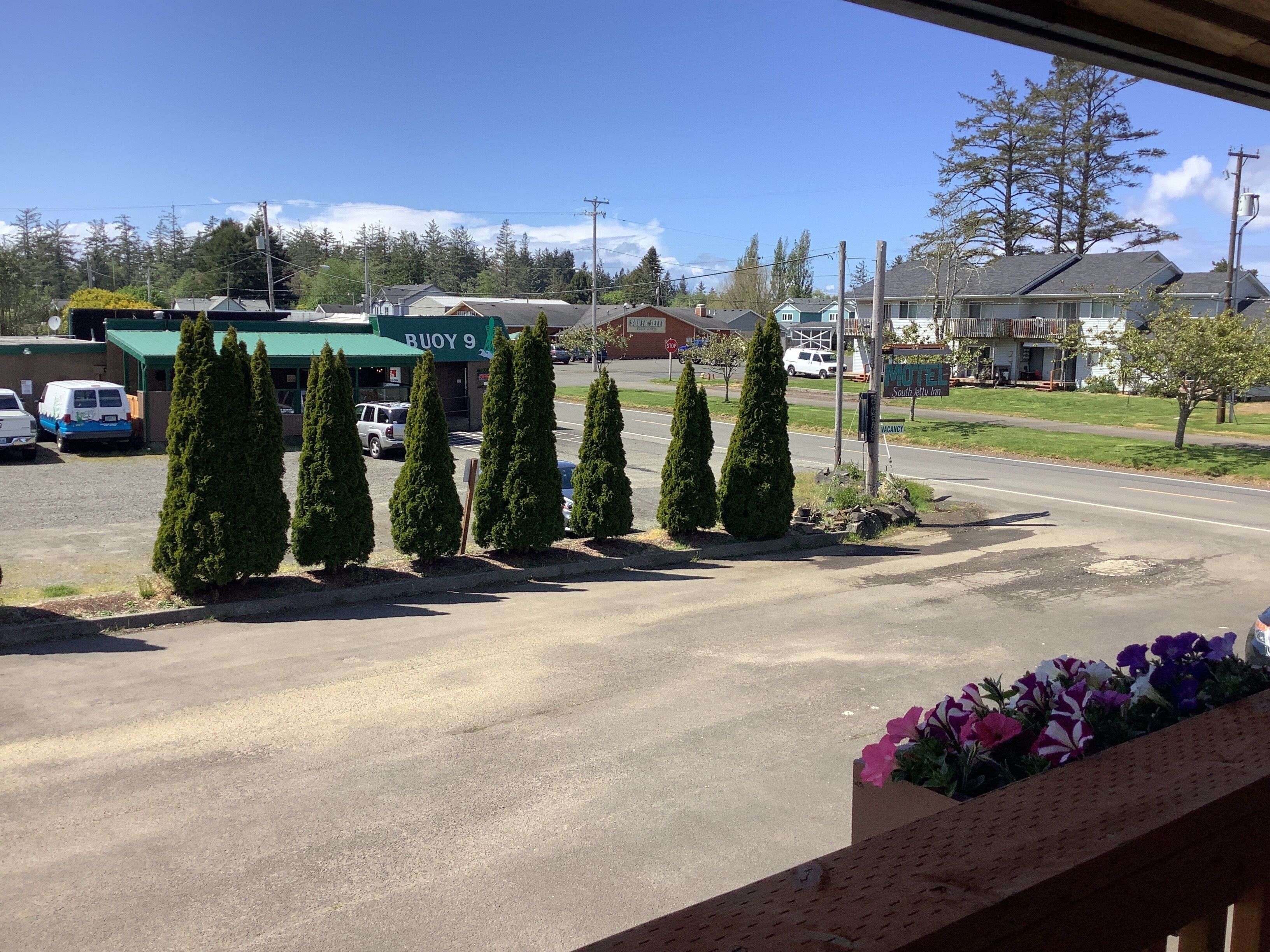 Un motel avec une enseigne verte, des véhicules garés et une rangée d’arbres à feuilles persistantes devant.