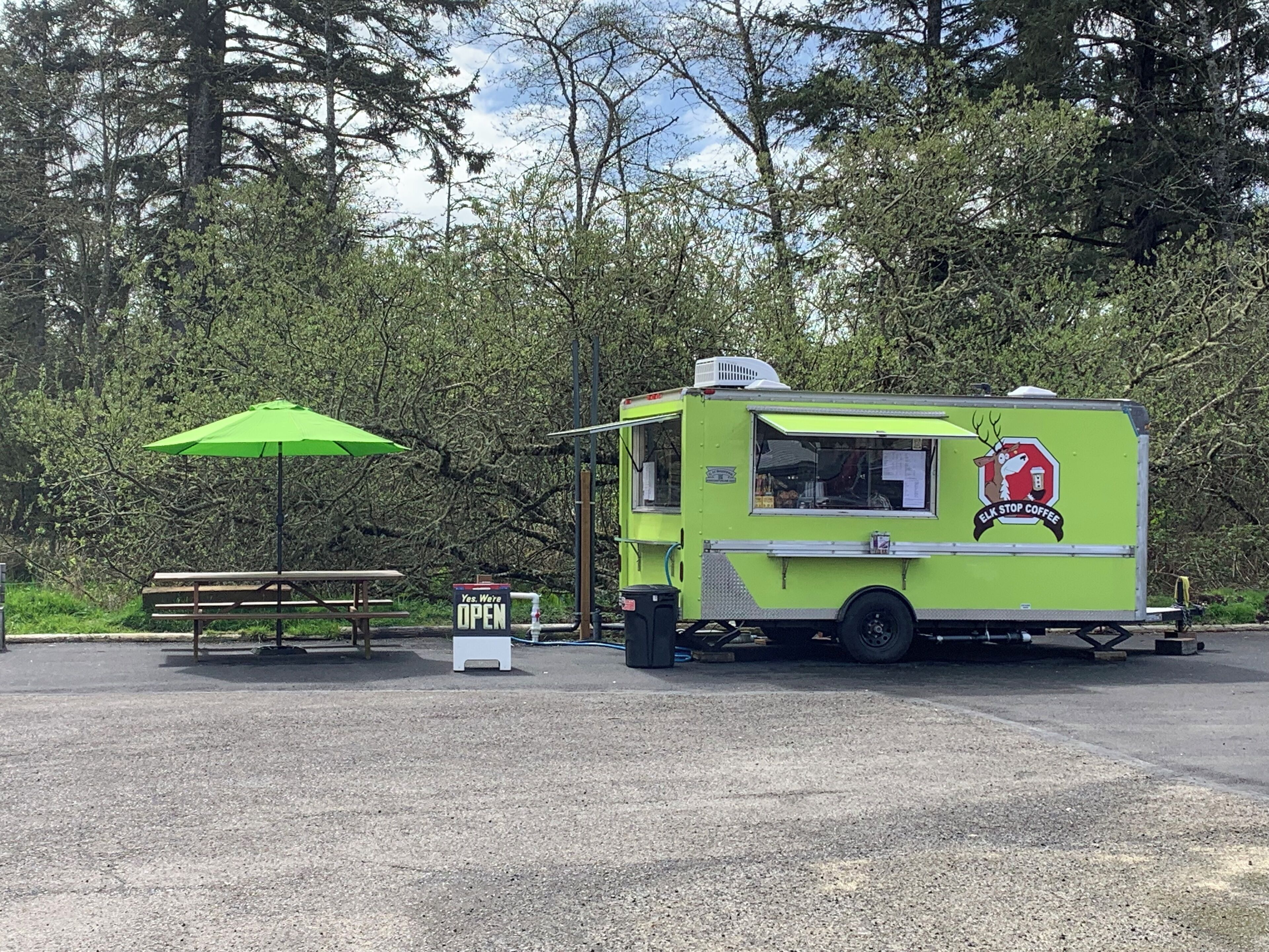 Un camion-restaurant vert, orné d’un logo, est garé sur un parking équipé d’une table de pique-nique et d’un parasol.
