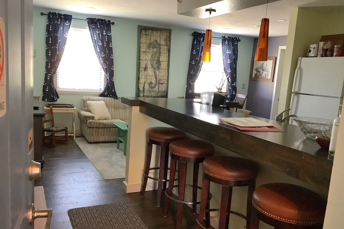 A kitchen with a bar area, wooden stools, a dining table, and a sofa.
