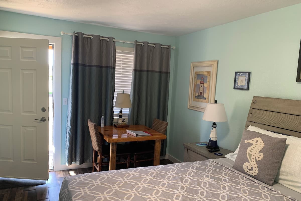 A bedroom with a bed, a wooden table, a chair, a nightstand, a lamp, and a picture on the wall.