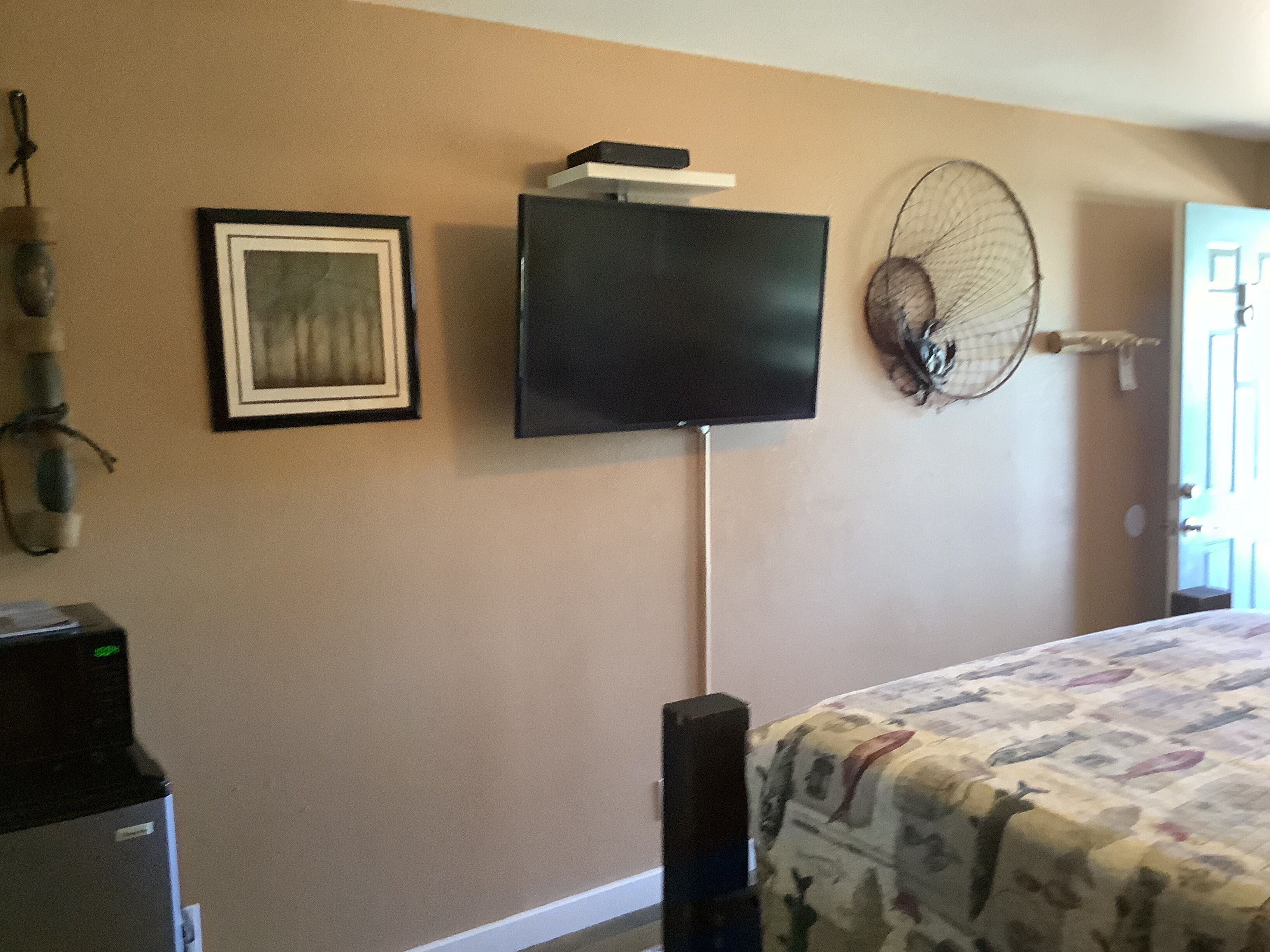 A bedroom with a bed, a television mounted on the wall, a framed picture, and a fan.