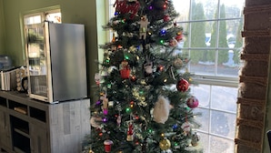 A Christmas tree decorated with ornaments and lights in a room with a window, a refrigerator, and a wooden cabinet. - South Jetty Inn (Hammond)
