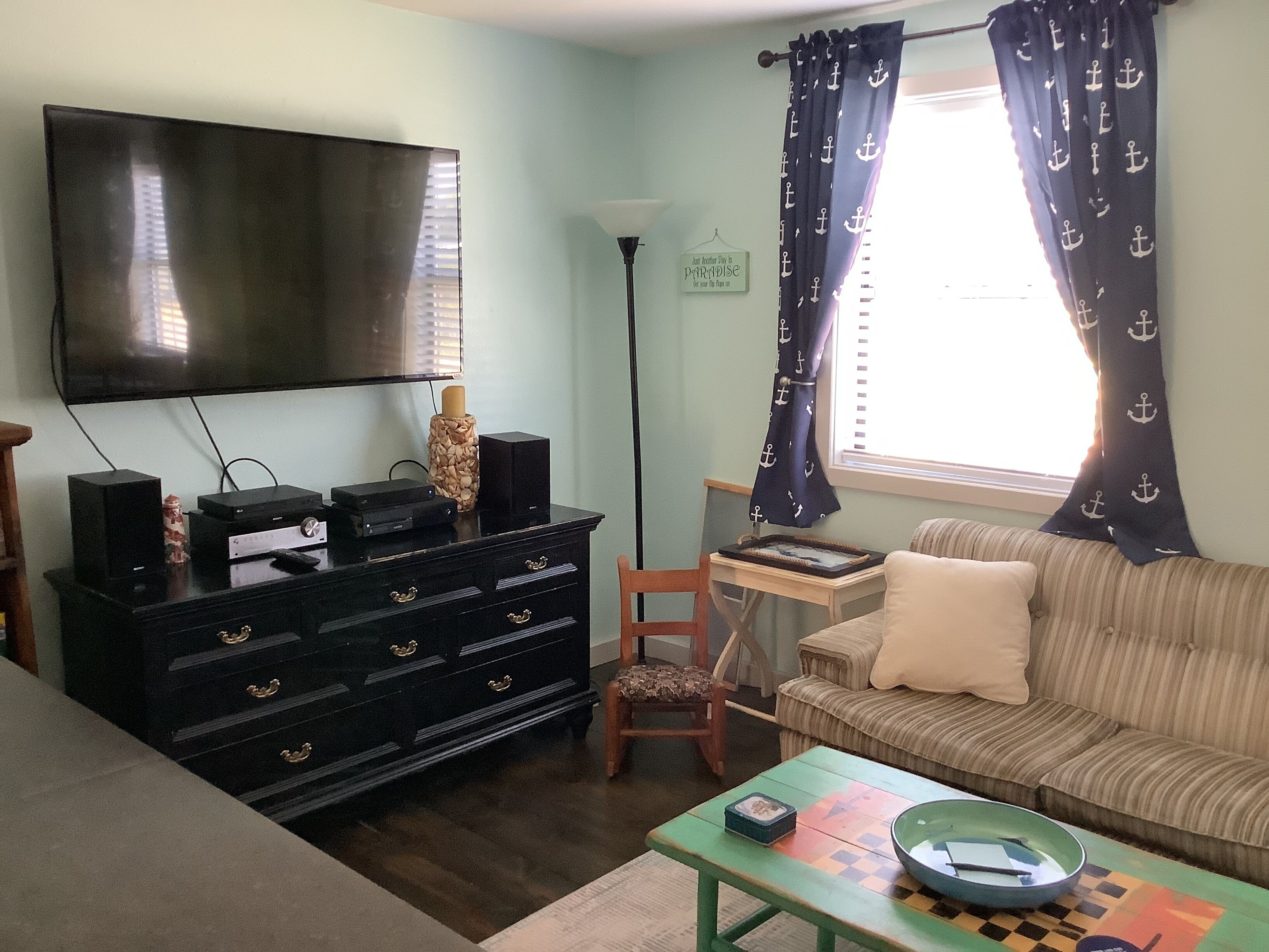 A living room with a flat-screen TV, a black entertainment unit, a patterned sofa, a wooden chair, and a small table with a bowl.