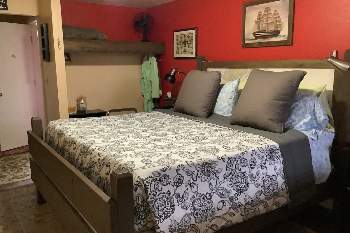 A bedroom with a bed, a nightstand, a lamp, a fan, and a picture on the wall.