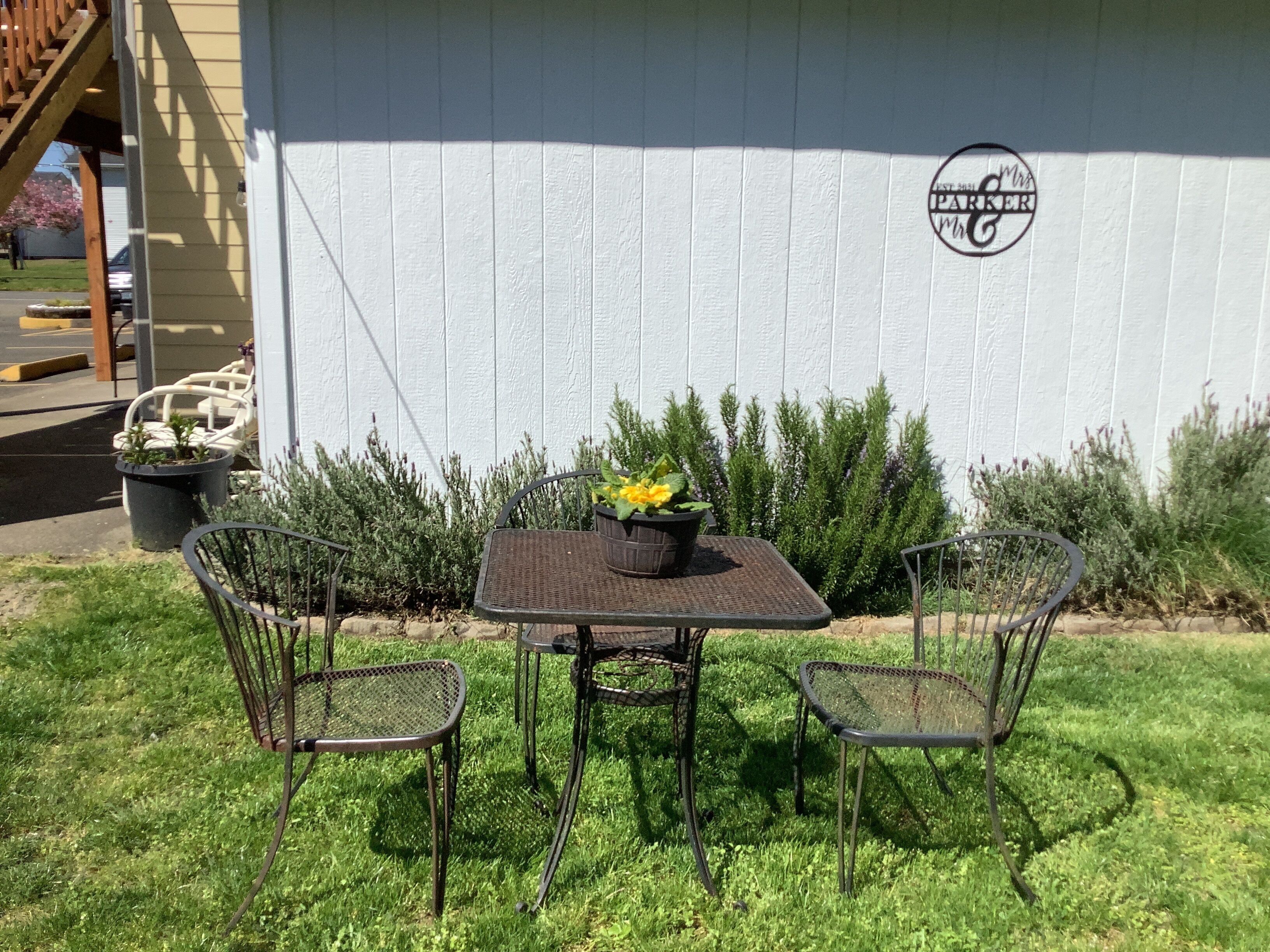 An outdoor seating area with a table and four chairs, a potted plant, and a white wall with a logo.