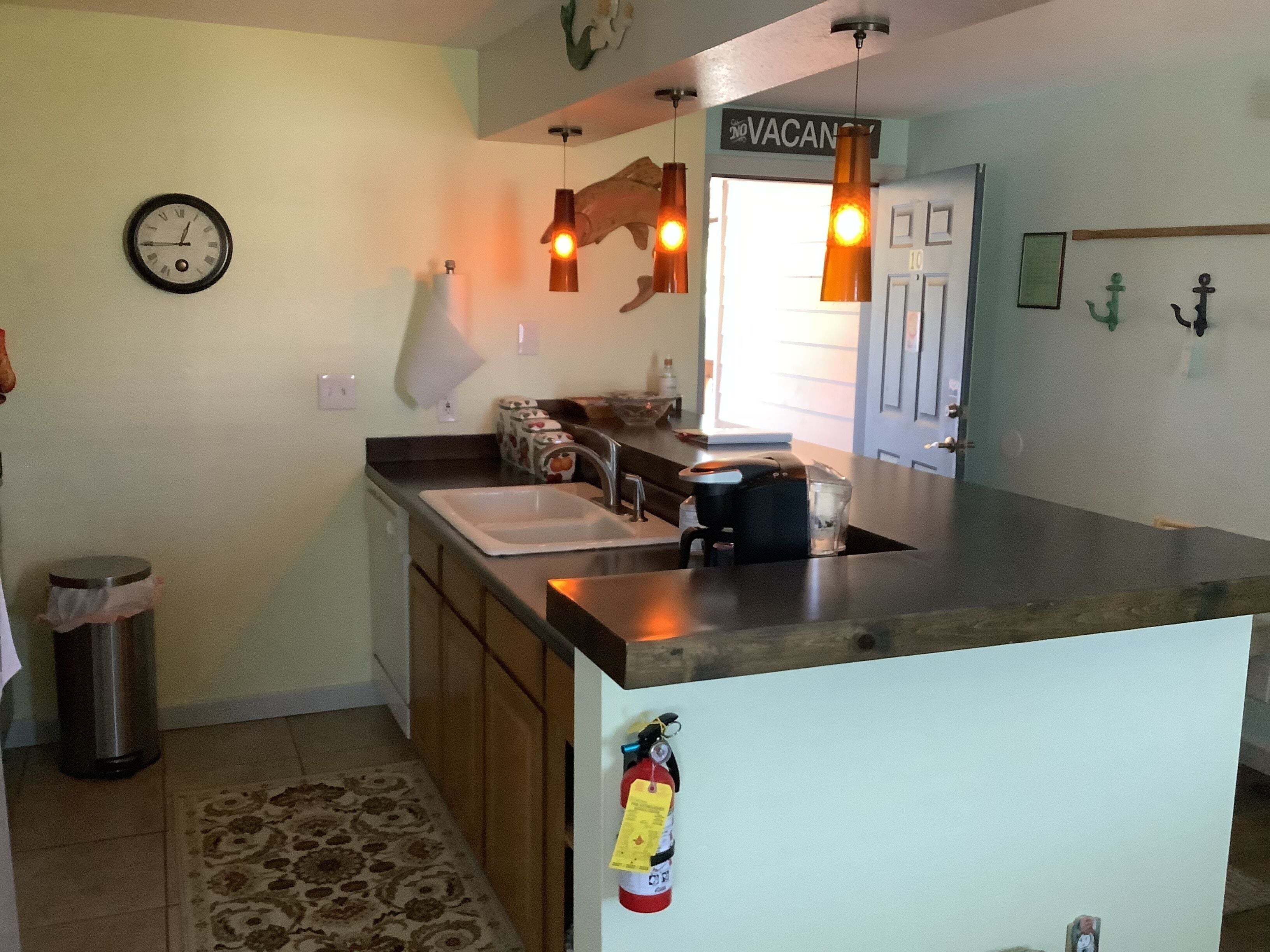 Una cocina con un lavamanos, cafetera y extintor. Hay un reloj en la pared y un letrero que dice "VACANCY".