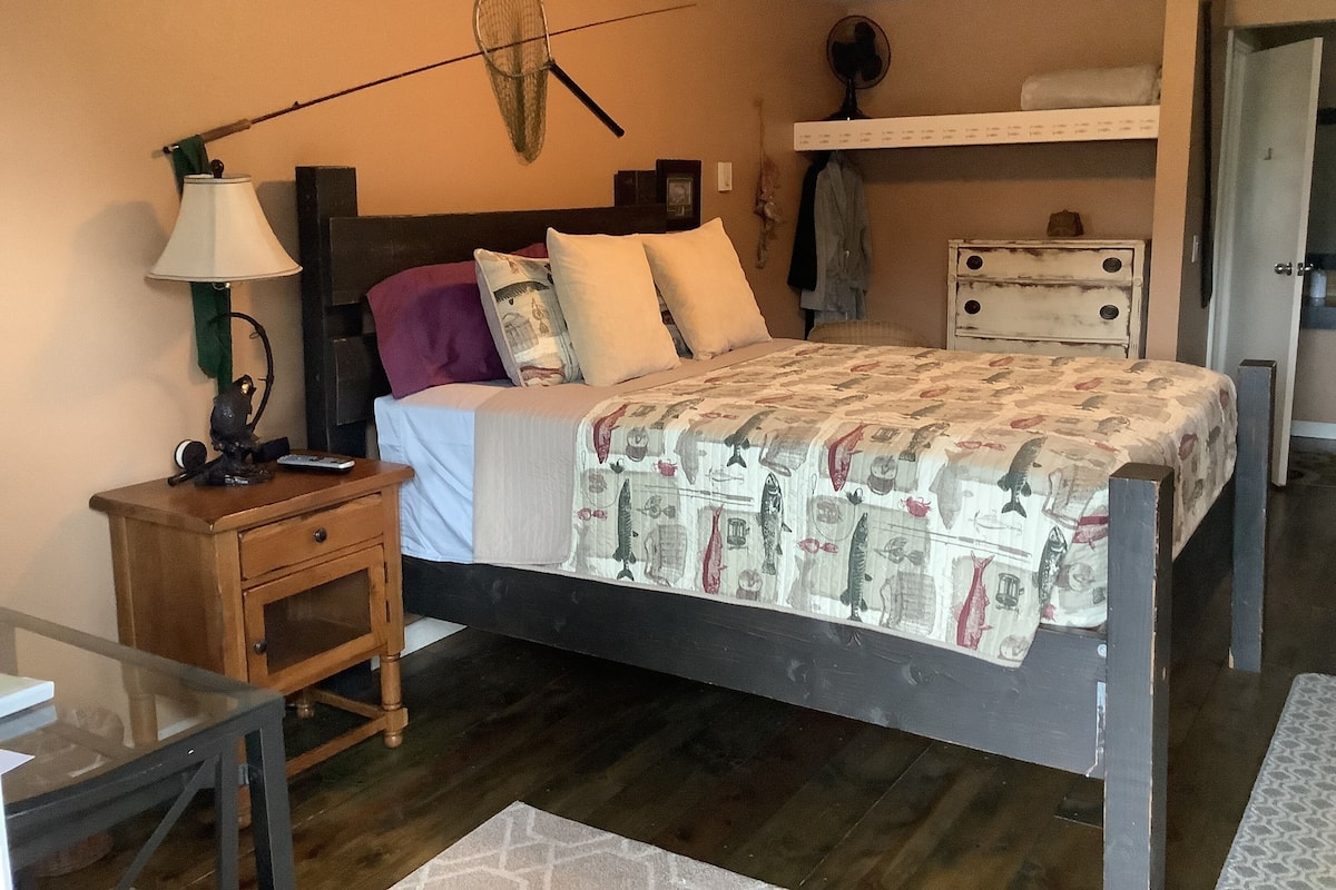 A bedroom with a bed, bedside tables, a lamp, a wall-mounted fan, and a shelf with clothes.