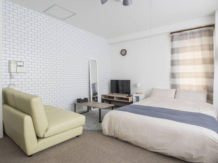 A modern hotel room with a white brick wall, a bed with a blue accent, a sofa, a TV, and a ceiling-mounted light fixture.
