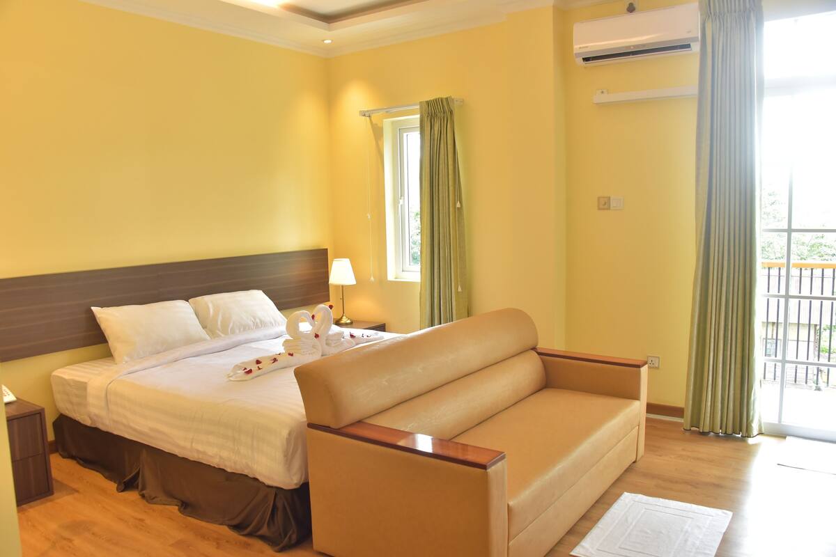 Een hotelkamer met een bed, een bank, een raam met gordijnen, een airconditioning en uitzicht op een balkon.