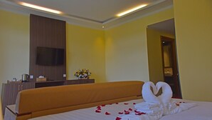 Habitación de hotel con una cama, un televisor montado en la pared, un aparador de madera y un florero con flores.