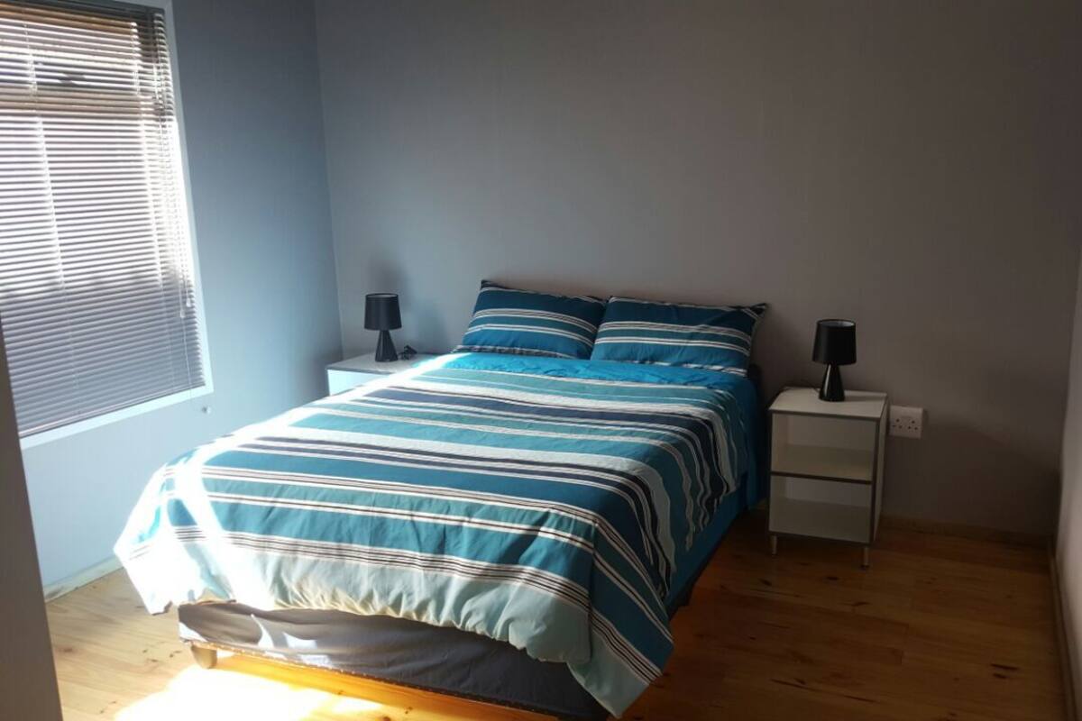 Un dormitorio con una cama, dos mesitas de noche, una ventana con persianas y una puerta.
