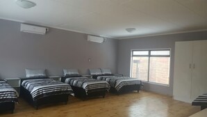Een kamer met zes bedden, gerangschikt in twee rijen, een groot raam met zonwering en een witte kast.