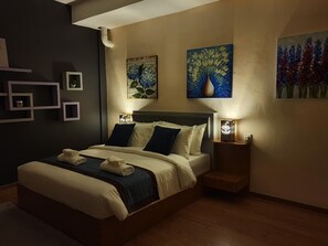 A bedroom with a large bed, two bedside tables with lamps, a wall-mounted painting, and a small shelf unit. - Assalam Hotel (Kota Bharu)