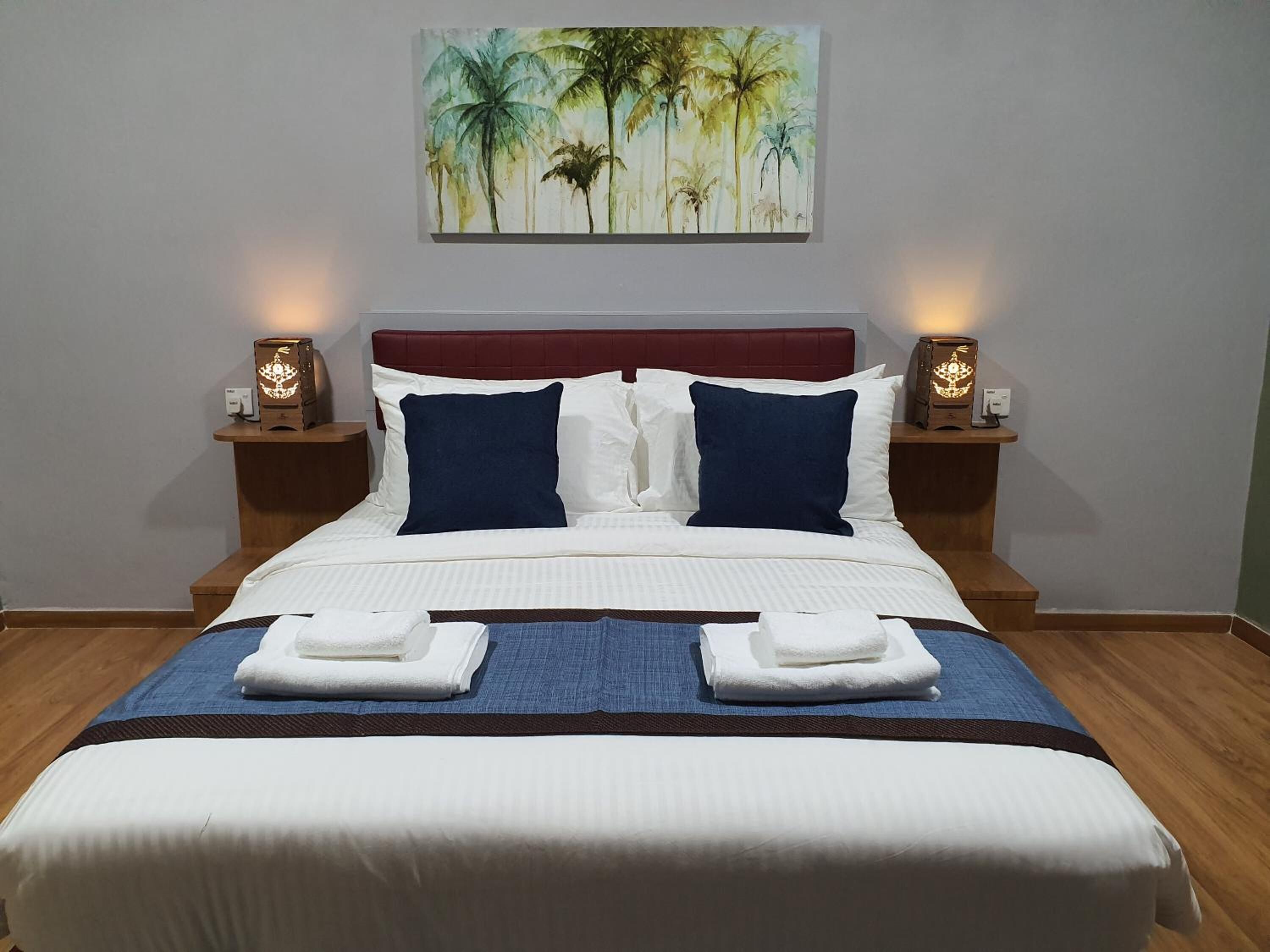 A neatly made bed with white linens, two blue pillows, and two white towels folded on the bed. There are two bedside tables with lamps, and a painting of palm trees on the wall above the bed.