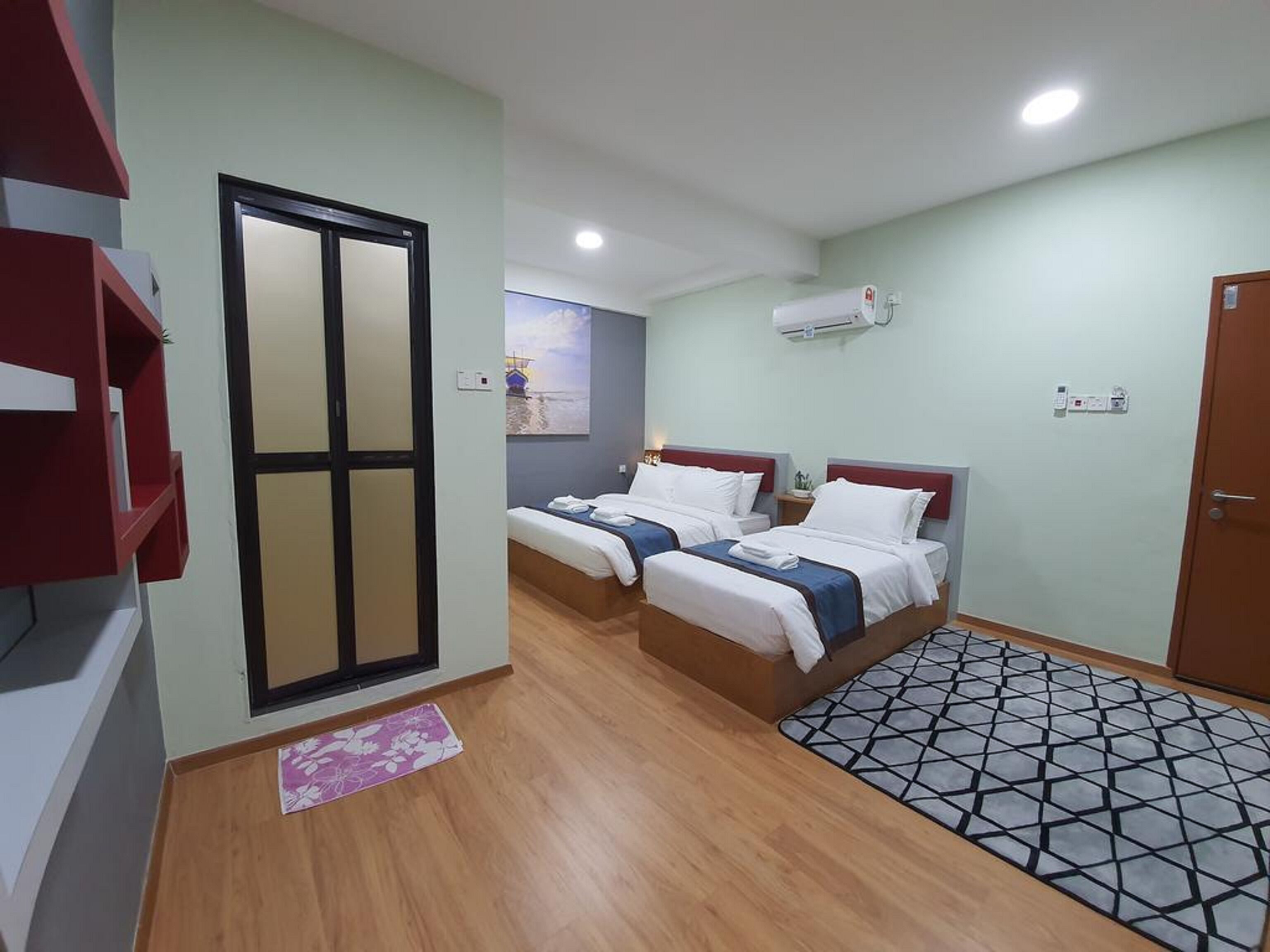 A hotel room with two beds, a wooden floor, a patterned rug, a door with a glass panel, and an air conditioning unit on the wall.