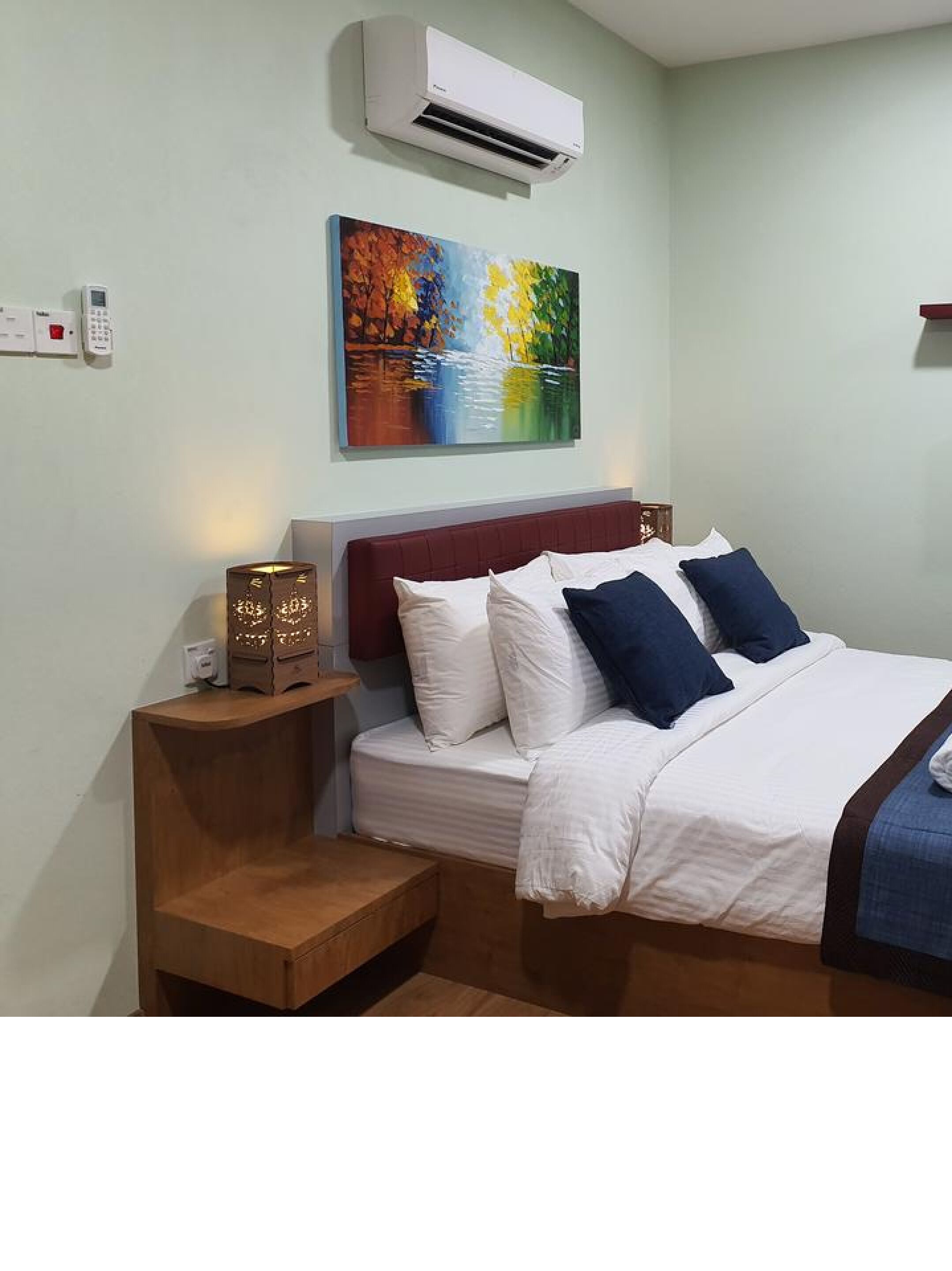 A hotel room with a bed, bedside table, nightstand, wall-mounted air conditioner, and a painting.