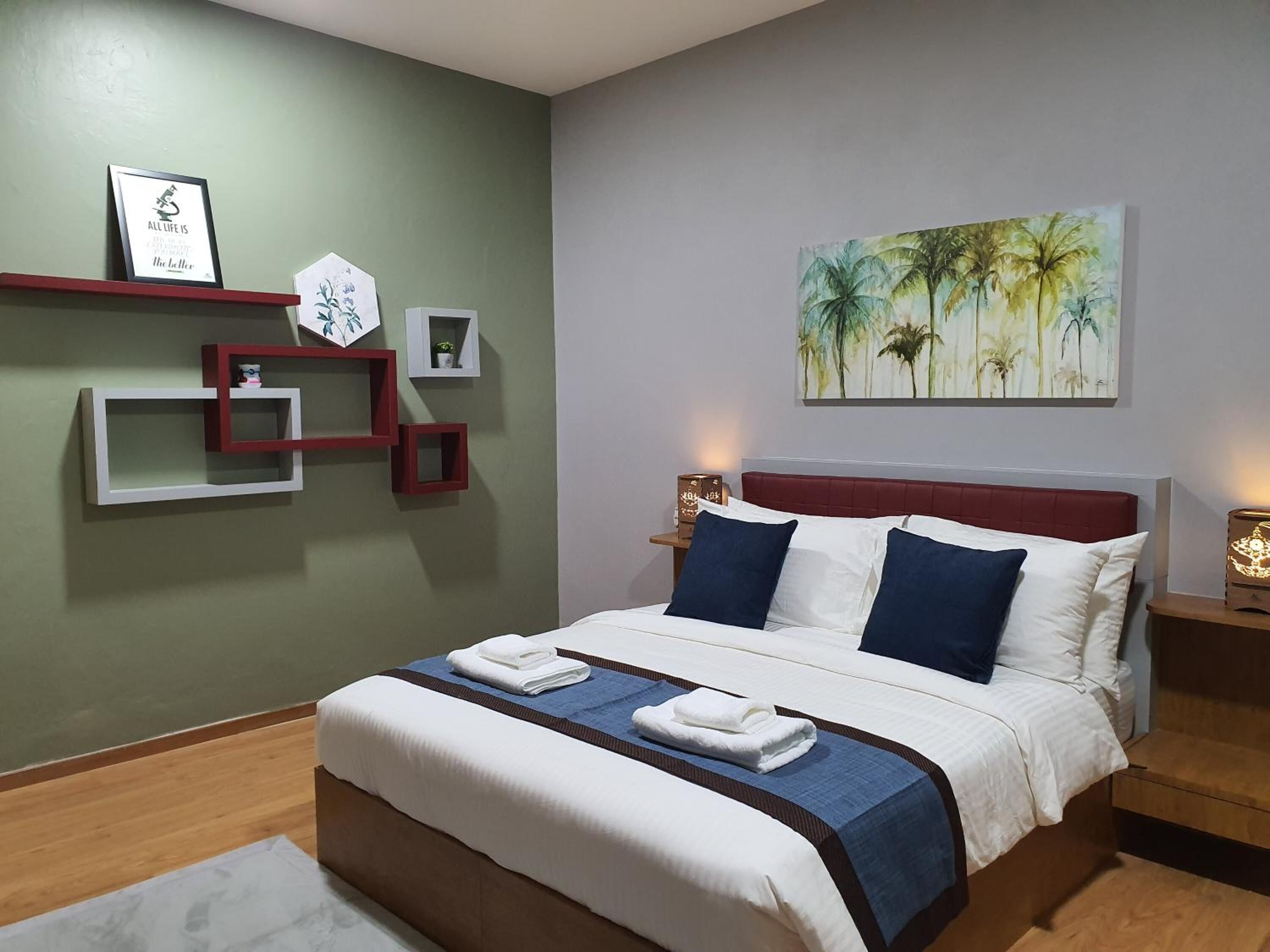 A neatly arranged hotel room with a bed, a wall-mounted shelf with decorative items, and a painting of palm trees.