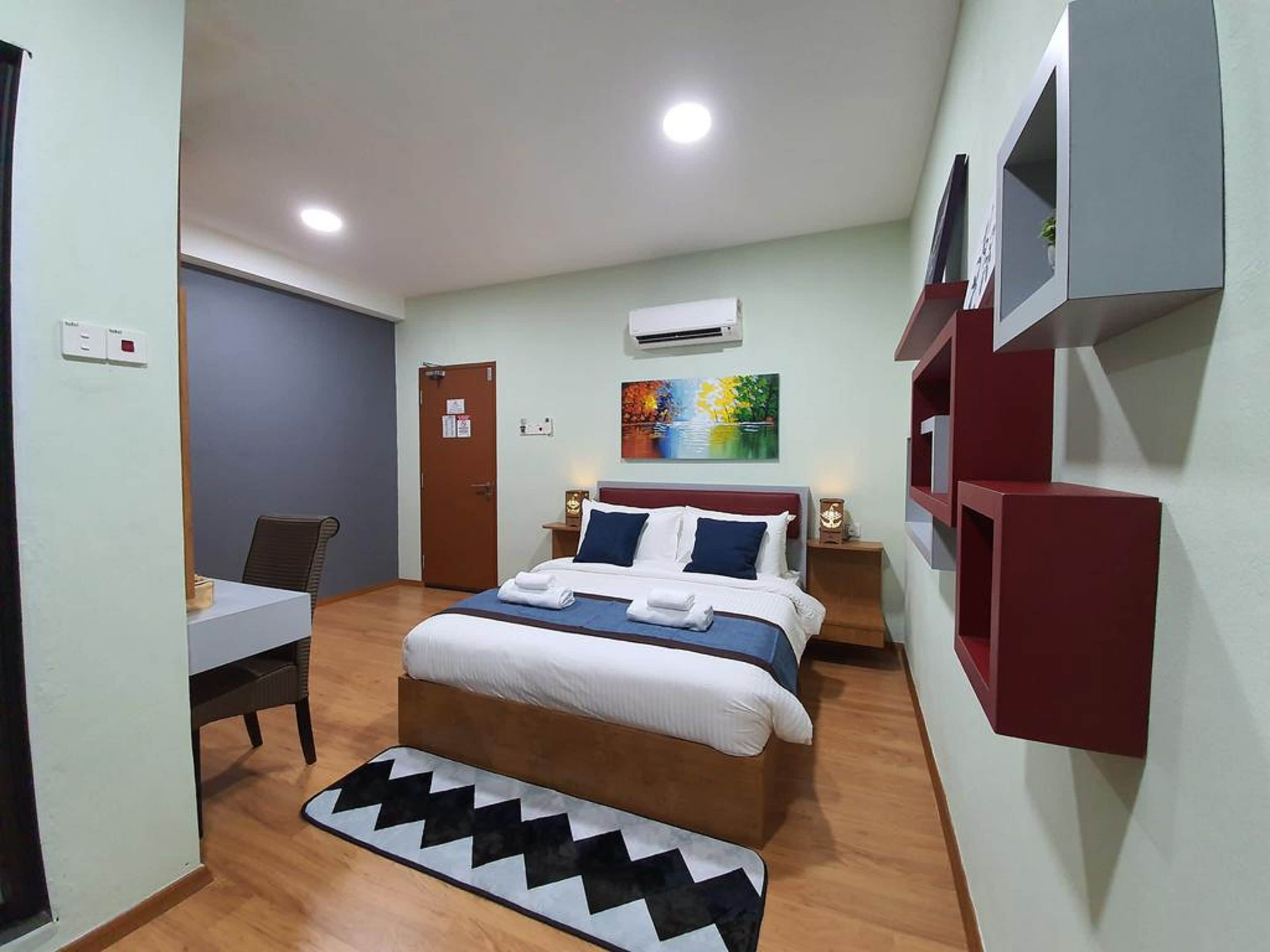 A hotel room with a bed, a desk, a chair, a small table, an air conditioning unit, and a painting on the wall.