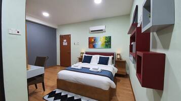 A hotel room with a bed, a desk, a chair, a small table, an air conditioning unit, and a painting on the wall.