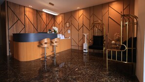 A reception area with a wooden reception desk, two bar stools, a standing lamp, and a decorative plant. - Assalam Hotel (Kota Bharu)