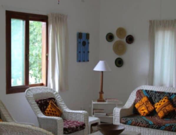 A living room with a sofa, armchair, and a table with a lamp. There are curtains on the windows and a wall decoration.