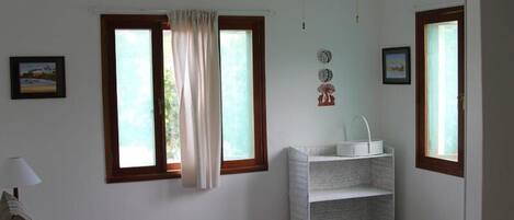 A bedroom with a bed, a bedside table, a lamp, a shelf, and a window with curtains.