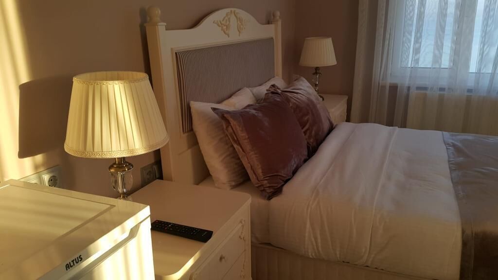 A neatly made bed with a headboard, two bedside lamps, a remote control on a nightstand, and a small drawer unit.