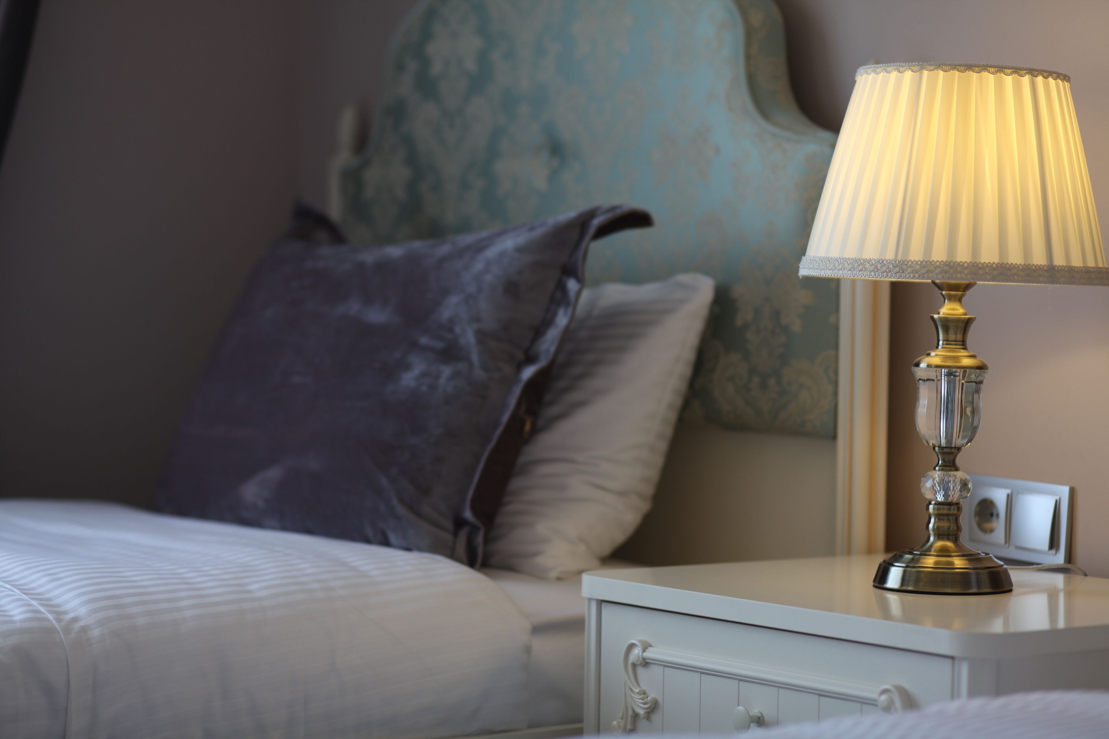 Ein ordentlich bezogenes Bett mit weißer Decke und Kissen, ein Nachttisch mit Lampe sowie eine Wand mit dekorativem, blauem Muster.