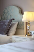 Una cama bien hecha con edredón blanco y almohadas, un cabecero tapizado en azul y una mesita de noche con lámpara.