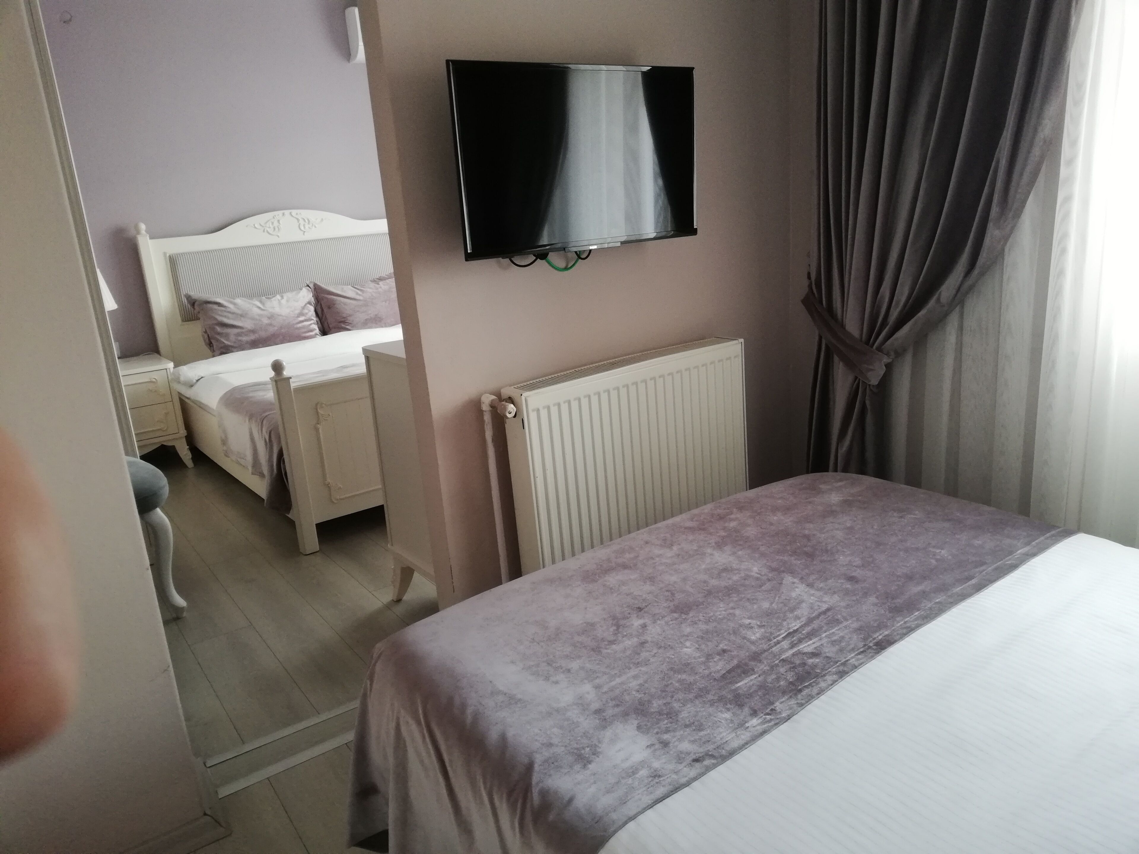 A bedroom with a bed, a TV mounted on the wall, a radiator, and a window with curtains.