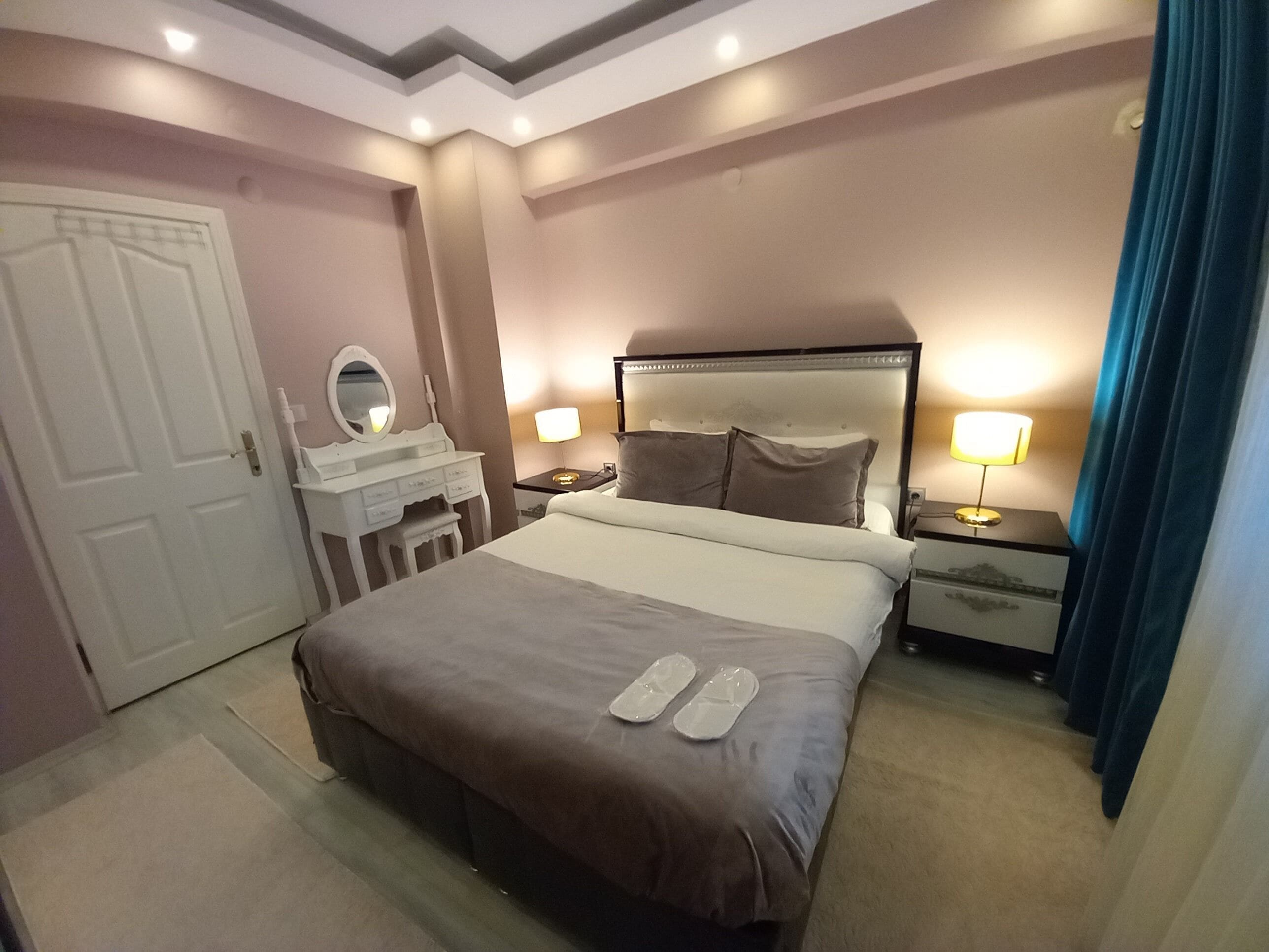 A hotel room with a large bed, a bedside table with a lamp, a dresser with a mirror, and a door.