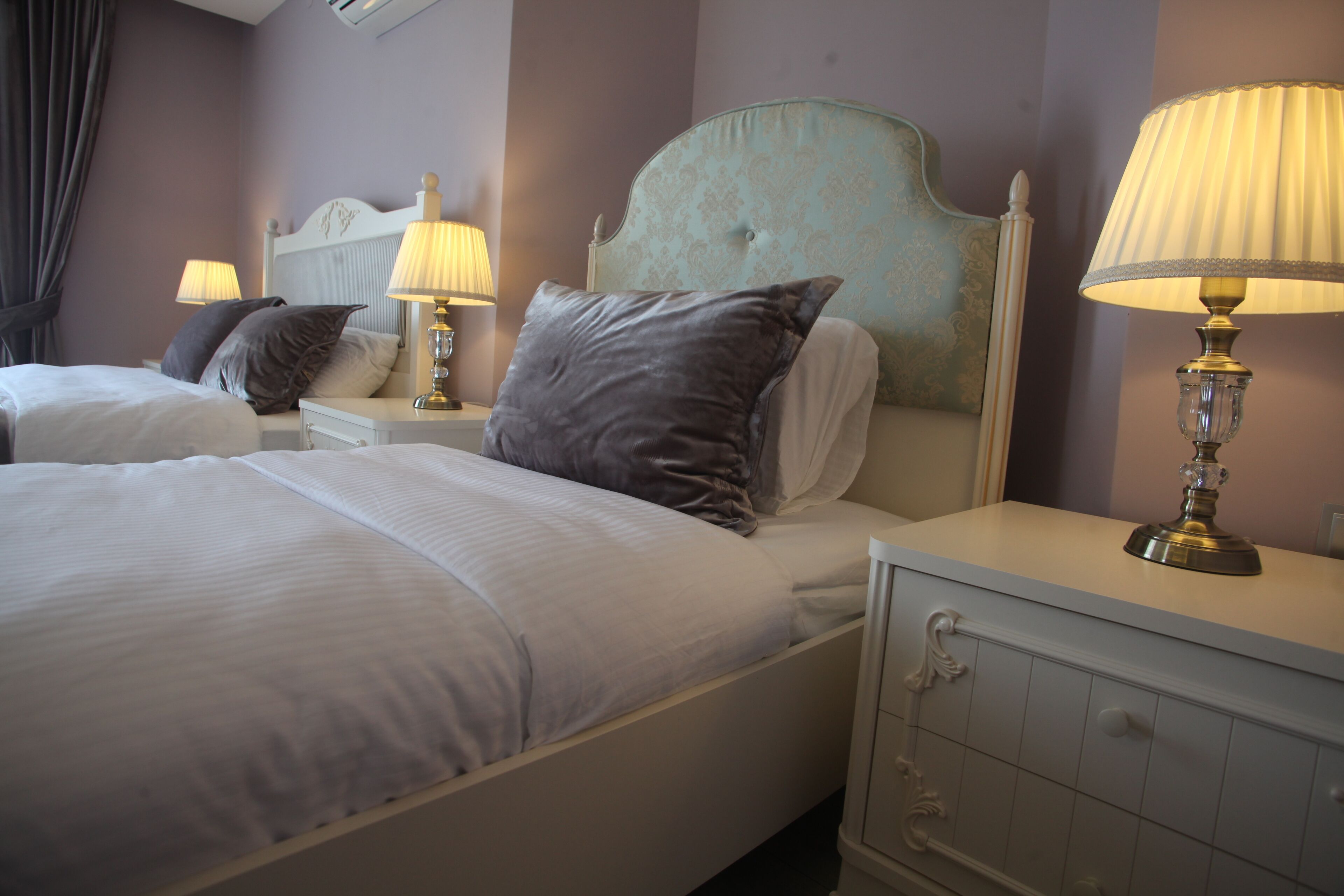 A hotel room with two beds, each with a headboard, bedside tables, lamps, and a dresser with drawers.