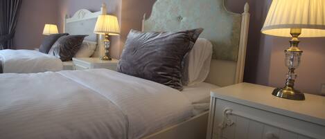 A hotel room with two beds, each with a headboard, bedside tables, lamps, and a dresser with drawers.