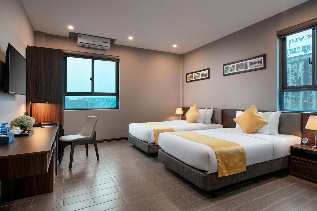 A hotel room with two beds, a desk, and a chair. There is a window with a view of the city.