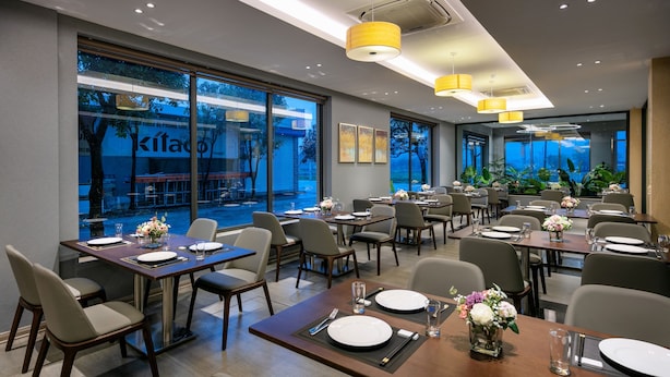 Un espace de restauration moderne, avec des tables dressées pour le service, de grandes fenêtres offrant une vue sur l’extérieur et un éclairage d’ambiance.