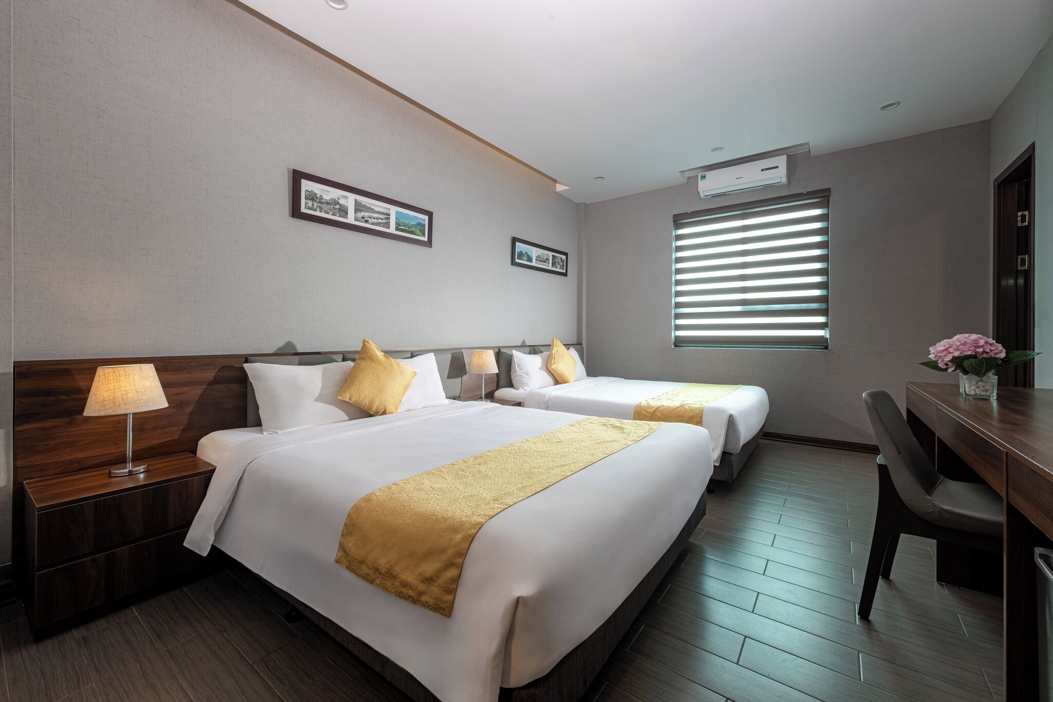 A hotel room with two beds, a desk, a chair, a lamp, and a window with blinds.