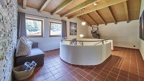 Una habitación con pared de piedra, techo de madera, bañera redonda, banca acolchada y ventana con cortinas.