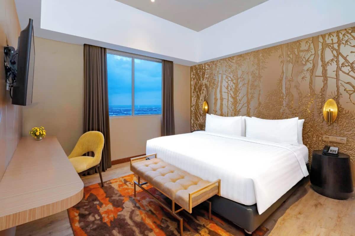 Habitación de hotel con una cama grande, un escritorio, una silla, un banco, un televisor y vistas a la ciudad.