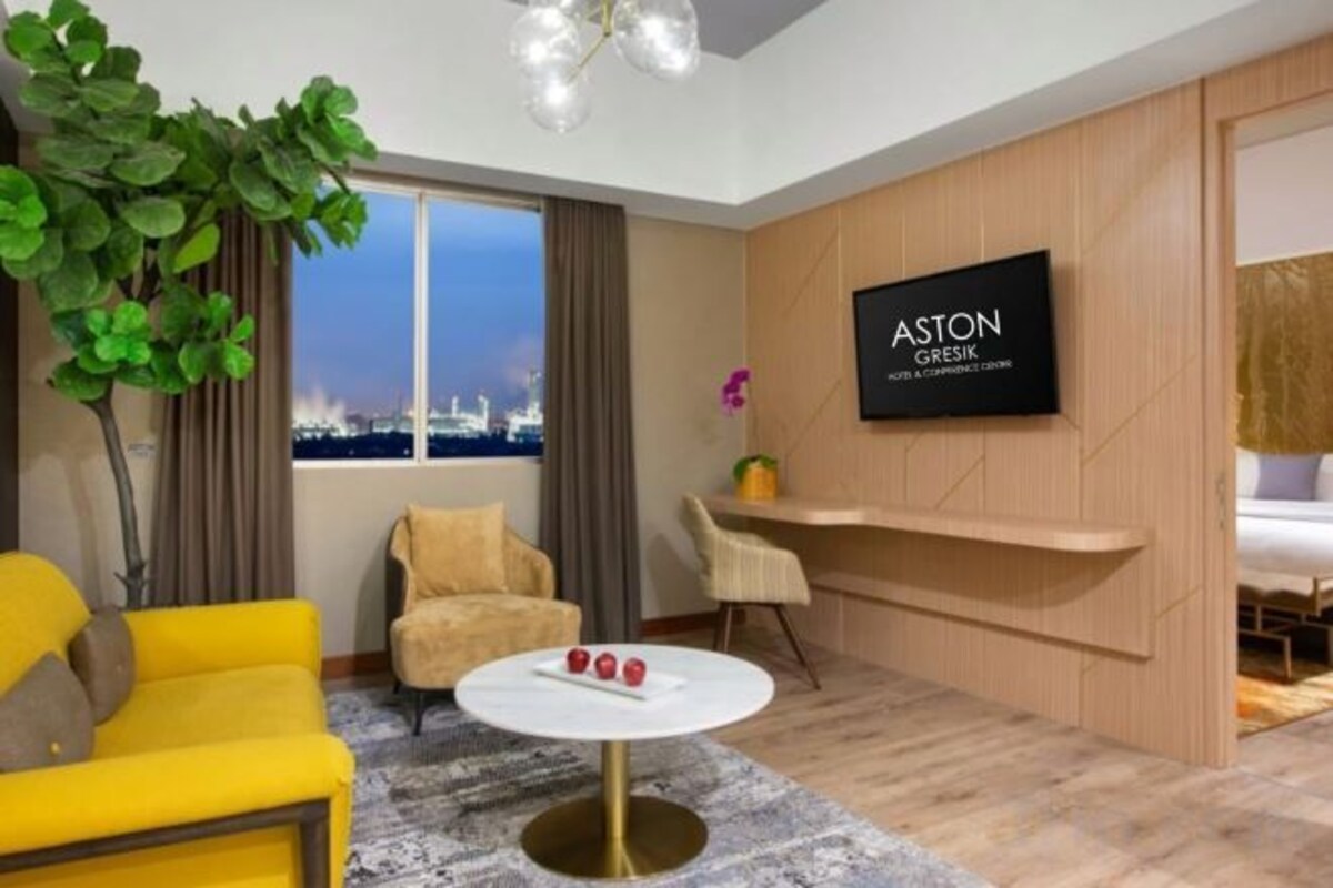 A modern hotel room with a city view, a flat-screen TV, a yellow sofa, a round coffee table, and a potted plant.