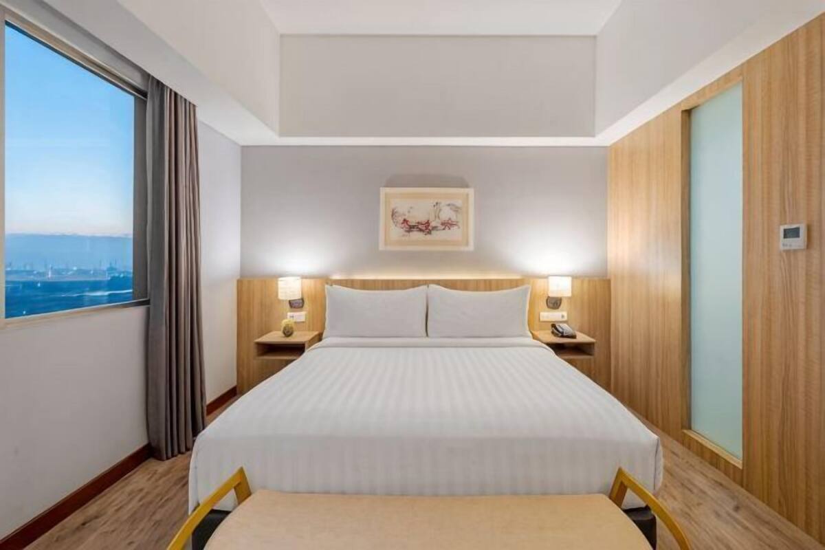 Habitación de hotel con una cama grande, dos mesitas de noche con lámparas, vista a la ciudad y un cabecero de madera.