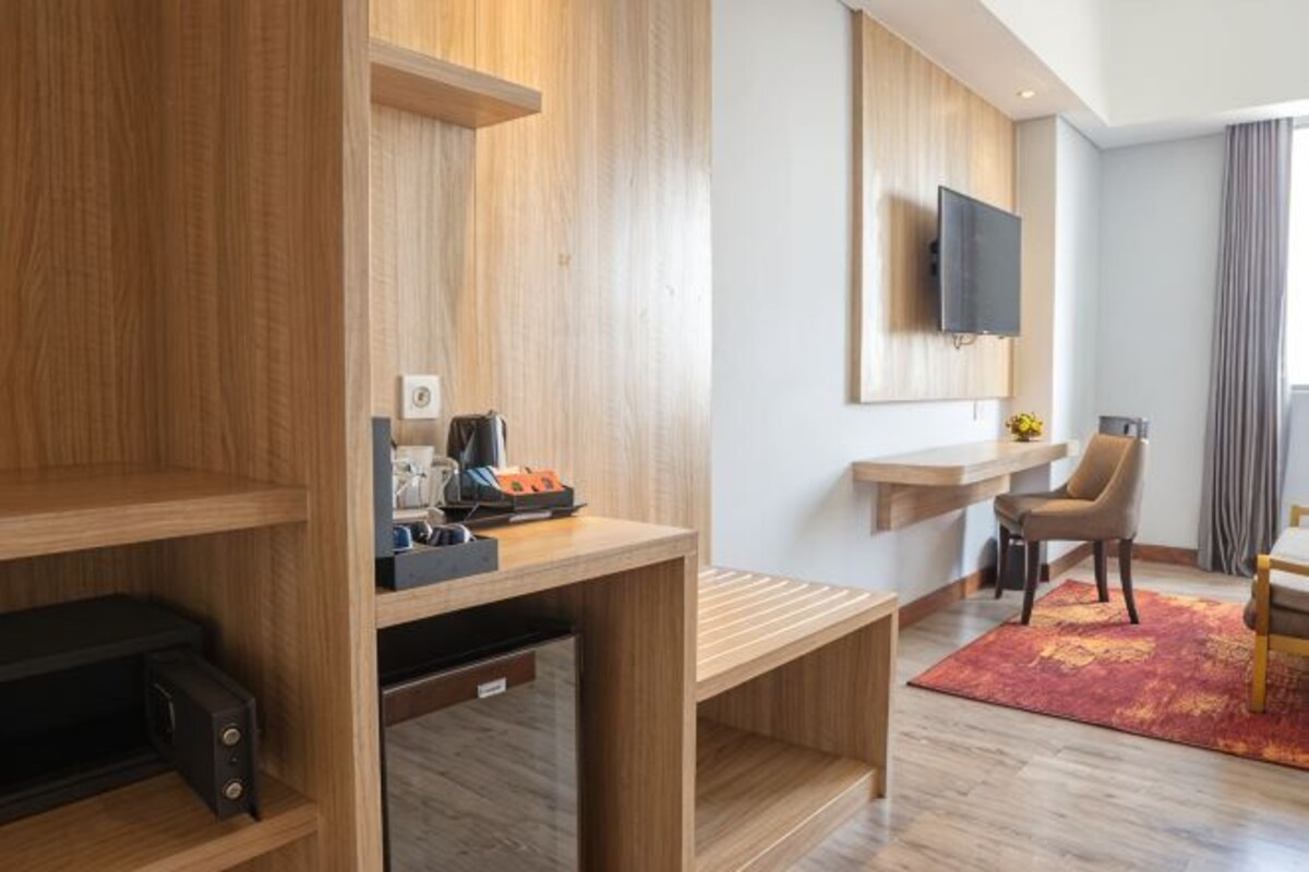 A hotel room with a wooden desk, a flat-screen TV mounted on the wall, a chair, and a small table.
