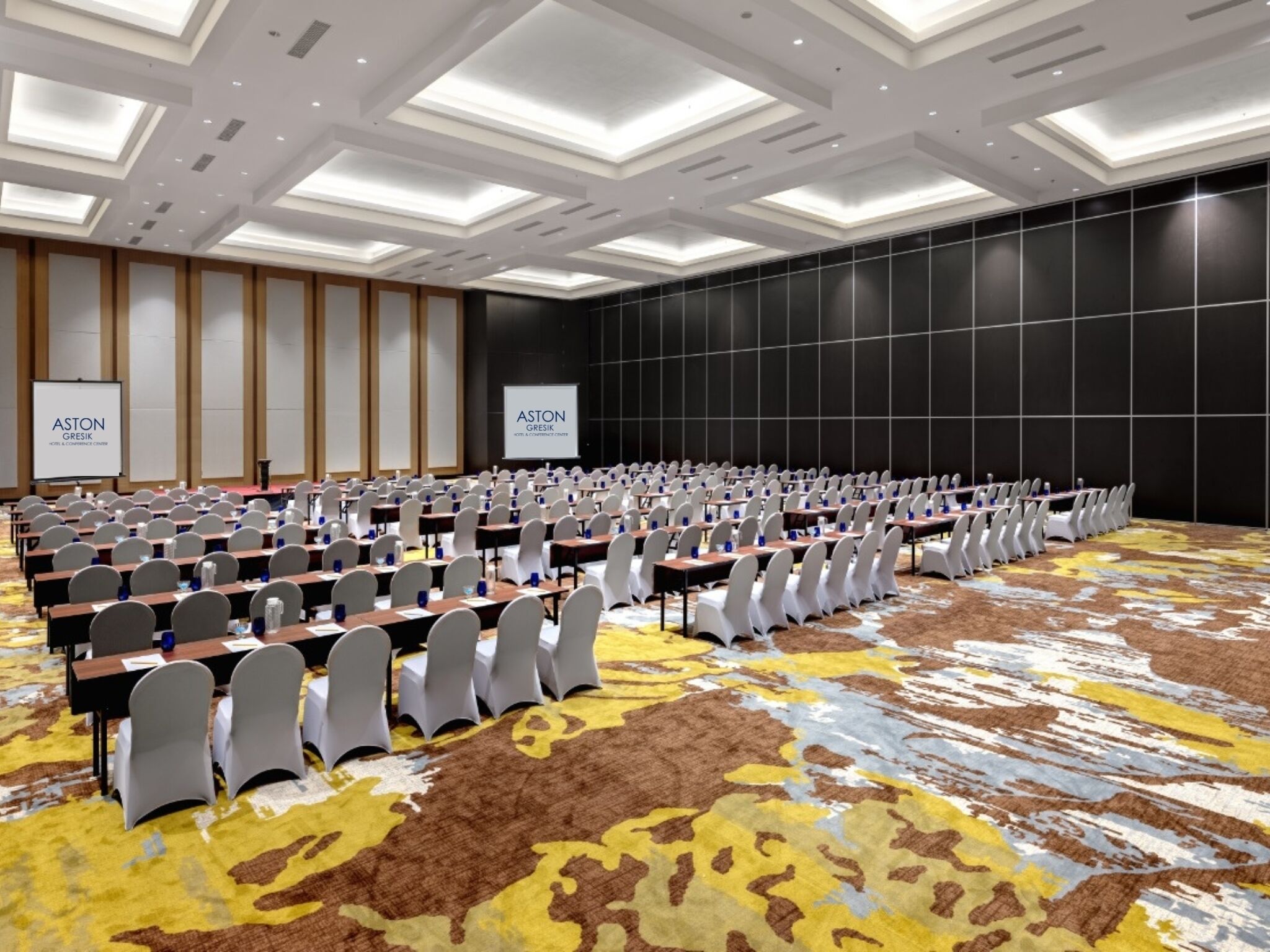 A conference room with rows of tables and chairs, a carpeted floor, and a large screen displaying the hotel name.