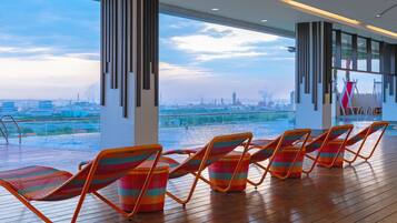 A modern indoor pool area with wooden flooring, colorful lounge chairs, and large windows offering a city view.