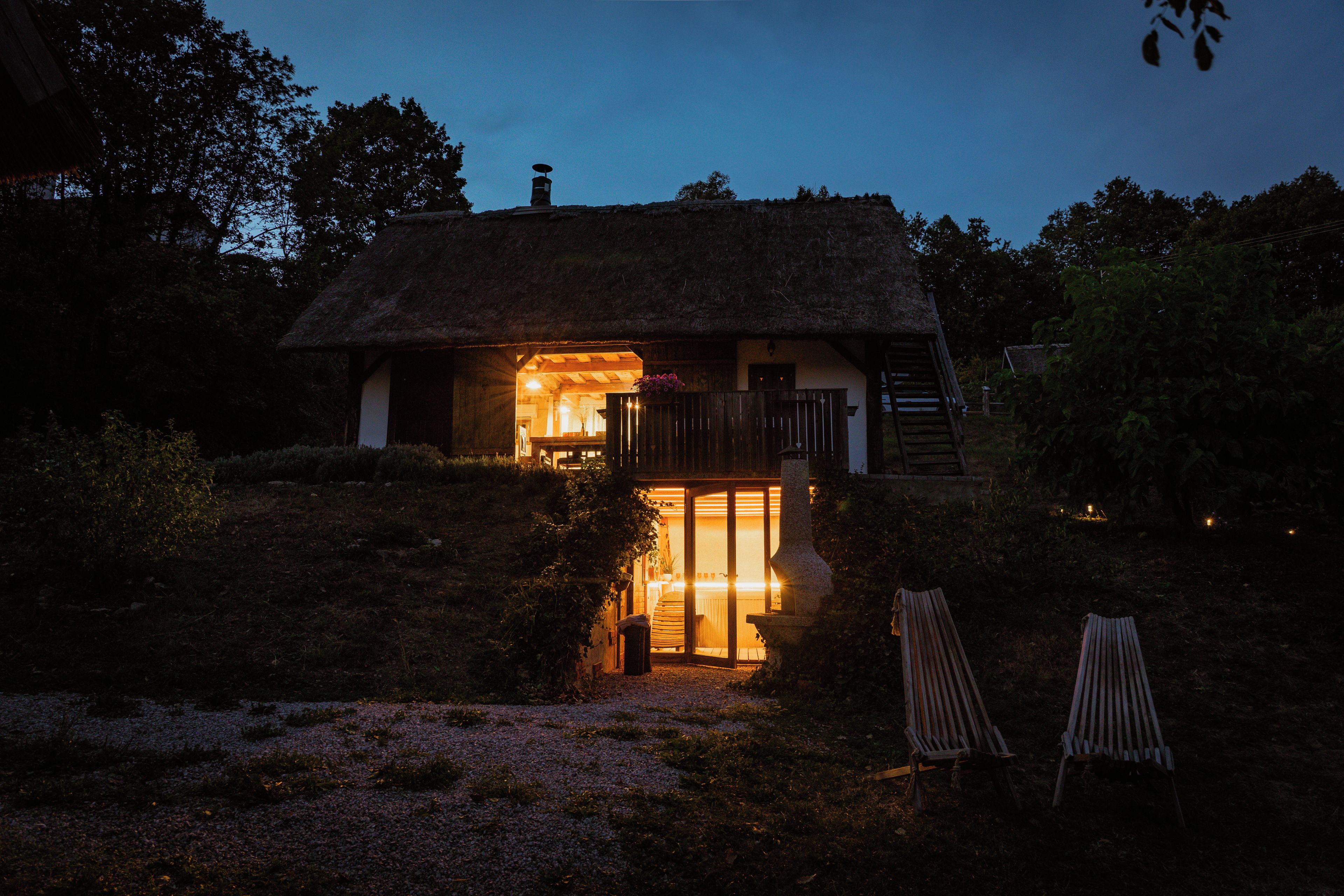 Ein reetgedecktes Cottage mit einem beleuchteten Innenraum, einer Holztür und zwei Holzstühlen davor.