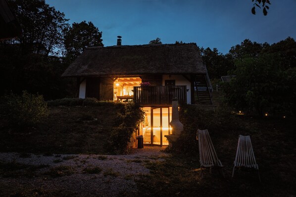 Une petite maison au toit de chaume, dont l’intérieur est éclairé, avec une porte en bois et deux chaises en bois à l’extérieur.